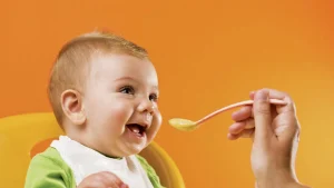 کودک نوزادی که لبخند زده و روی صندلی نشسته و فردی با قاشق به او غذا می دهد