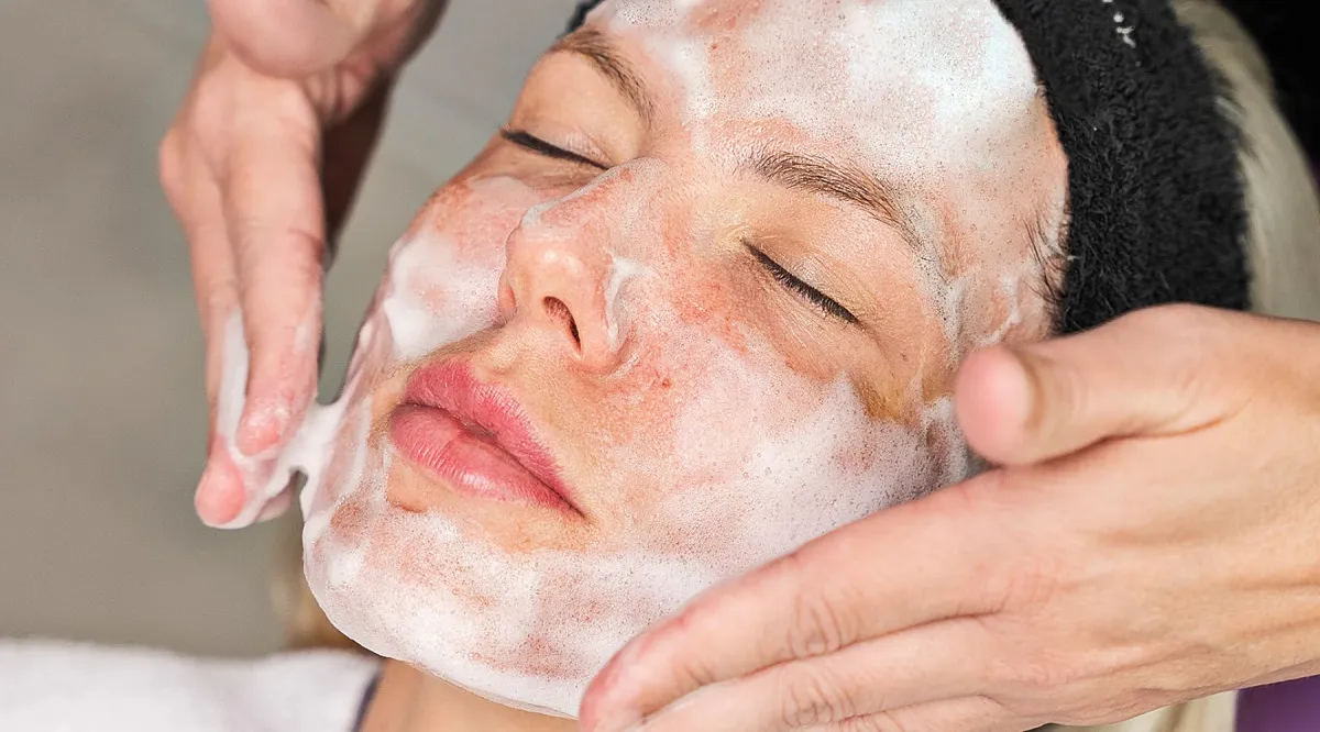 دختری که چشم خود را بسته است و فرد دیگری روی صورت او مواد فیشیال مالیده و صورت او را ماساژ می دهد