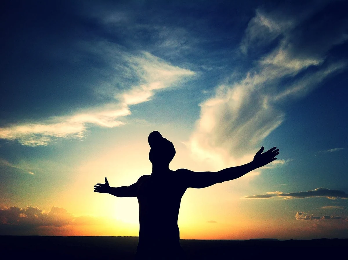 عکس از مردی که در فضای باز، جلوی نور خورشید ایستاده و دستان خود را باز کرده است