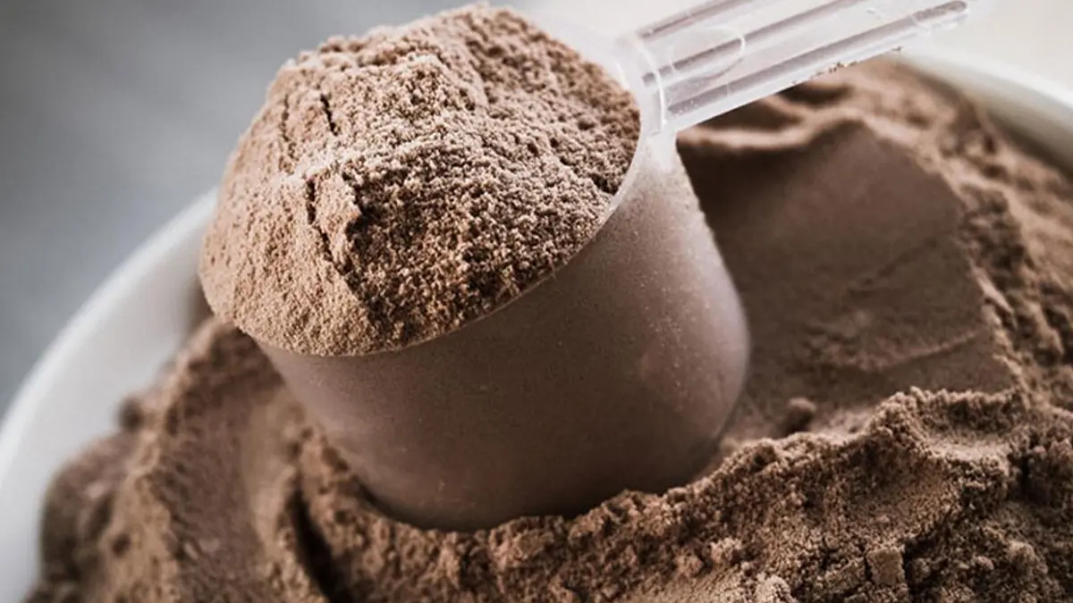 ظرف حاوی پودر مکمل بدنسازی قهوه ای رنگ که یک قاشق پر از پودر روی ظرف قرار گرفته