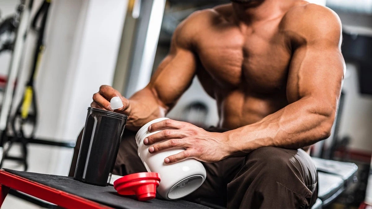 مرد بدنسازی که داخل باشگاه است و قوطی مکمل بدنسازی را در دست خود دارد و از پودر مکمل داخل لیوان خود خالی می کند
