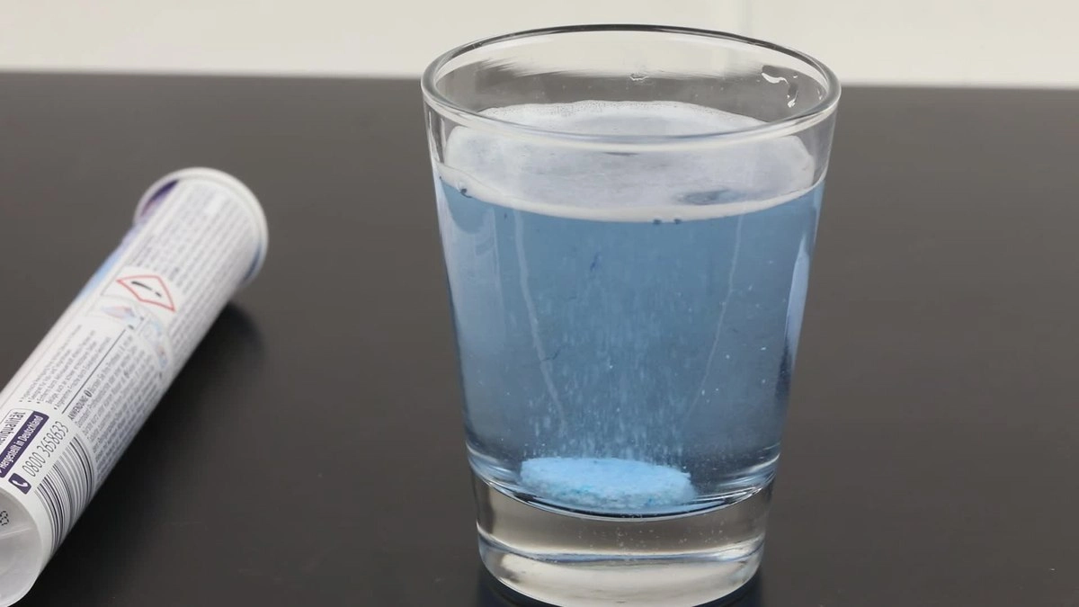 یک لیوان روی میز که یک قرص جوشان داخل آب آن در حال حل شدن است و قوطی قرص جوشان در کنار لیوان قرار گرفته است