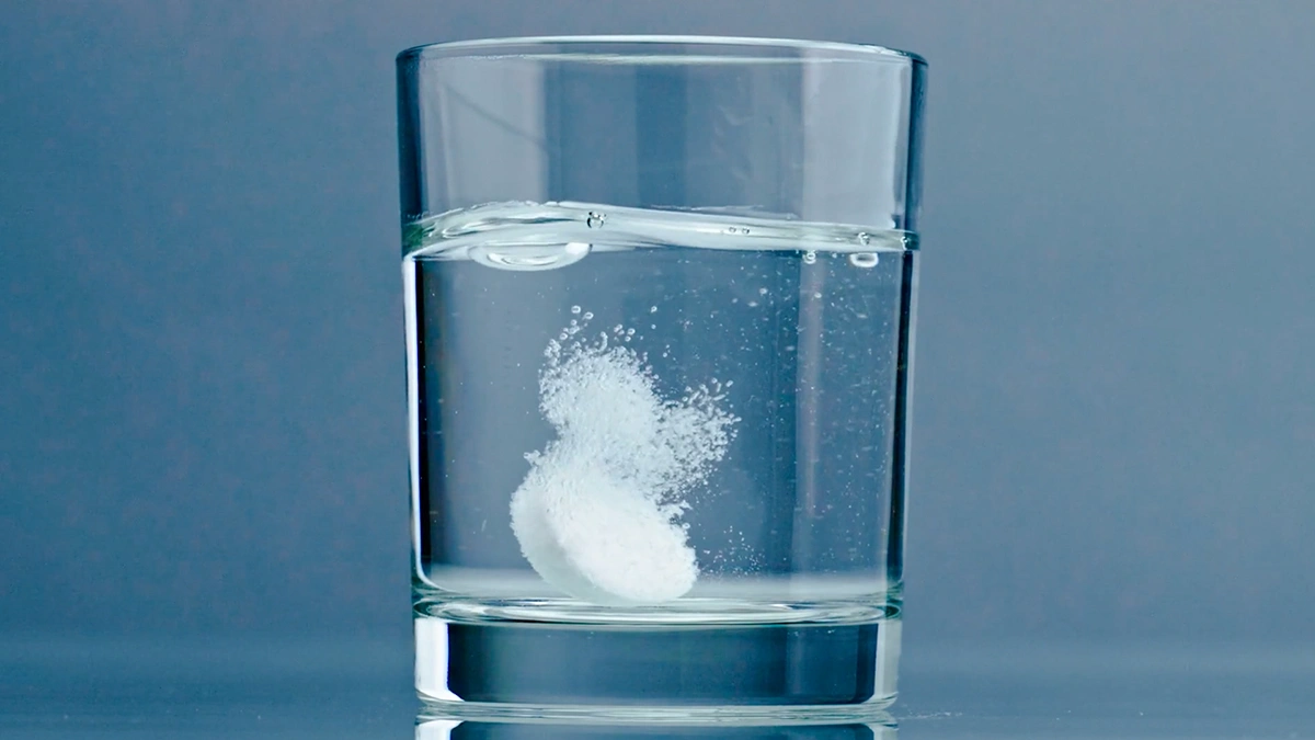 یک لیوان شیشه ای که درون آن آب است و یک قرص جوشان در حال حل شدن است