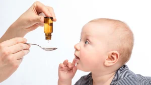 دستان فرد بزرگسال که قطره تقویتی کودک را داخل قاشق خالی می کند و نوزادی که به او نگاه می کند