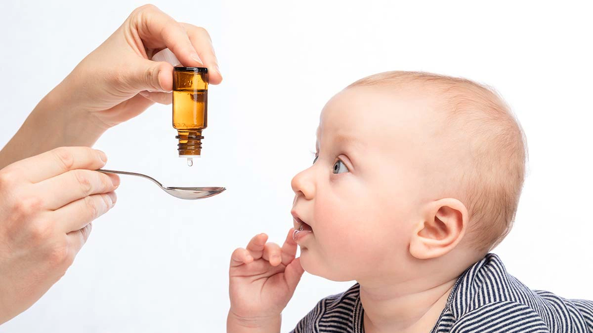 دستان فرد بزرگسال که قطره تقویتی کودک را داخل قاشق خالی می کند و نوزادی که به او نگاه می کند