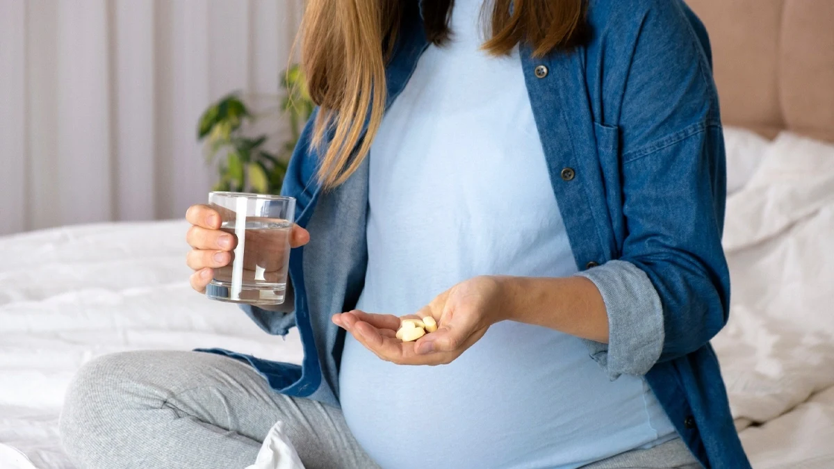 زن باردار که در یک دست قرص های مکمل بارداری و در دست دیگر یک لیوان آب دارد