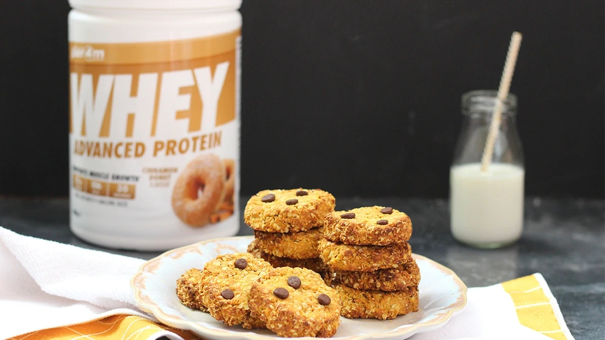 عکس یک بشقاب پر از کوکی که در کنار آن یک قوطی پروتئین وی و در سمت دیگر یک شیشه شیر قرار گرفته است